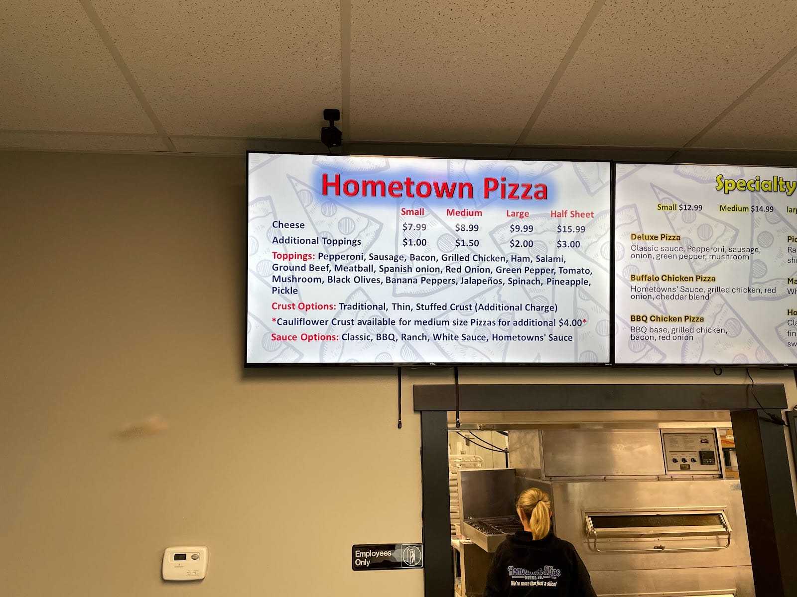 Digital menu board displaying options and prices for Hometown Pizza, with an employee in the background.